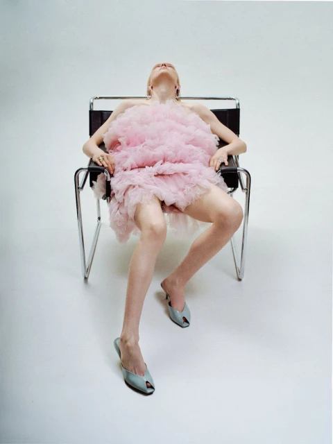 A young adult female is seated in a modern chair in a photo studio, wearing a pink dress and stylish shoes. Full body shot.