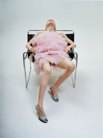 A young adult female is seated in a modern chair in a photo studio, wearing a pink dress and stylish shoes. Full body shot.