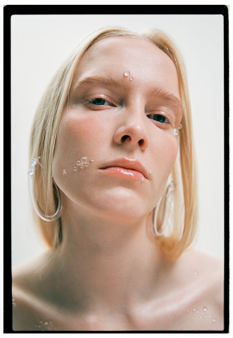 Close-up beauty shot of a young adult woman with blonde hair and water droplet makeup, wearing clear hoop earrings against white backdrop.
