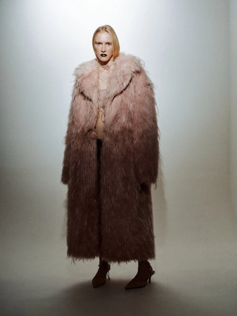 Fashion portrait of a blonde woman in a long pink faux fur coat and tan heels, standing in a photo studio against a neutral backdrop.