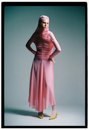 Full shot of a female fashion model in a pink head covering, long-sleeve ruched top, and skirt, posing indoors with hands on hips.