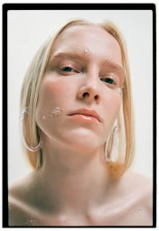 Close-up portrait of a blonde model with water droplets on her face and clear hoop earrings against a white background