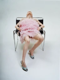 Model wearing a voluminous pink ruffled dress seated in a modern chrome framed leather chair against a white background.