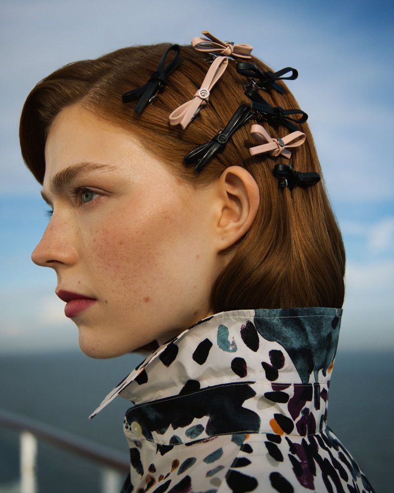 Close-up of a woman's profile with auburn hair styled with decorative hair bows, wearing a patterned collared shirt against a sky backdrop.