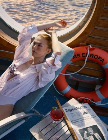 Woman relaxes on boat deck in sunlight wearing an open shirt; water visible through porthole, accompanied by cocktail and newspaper.