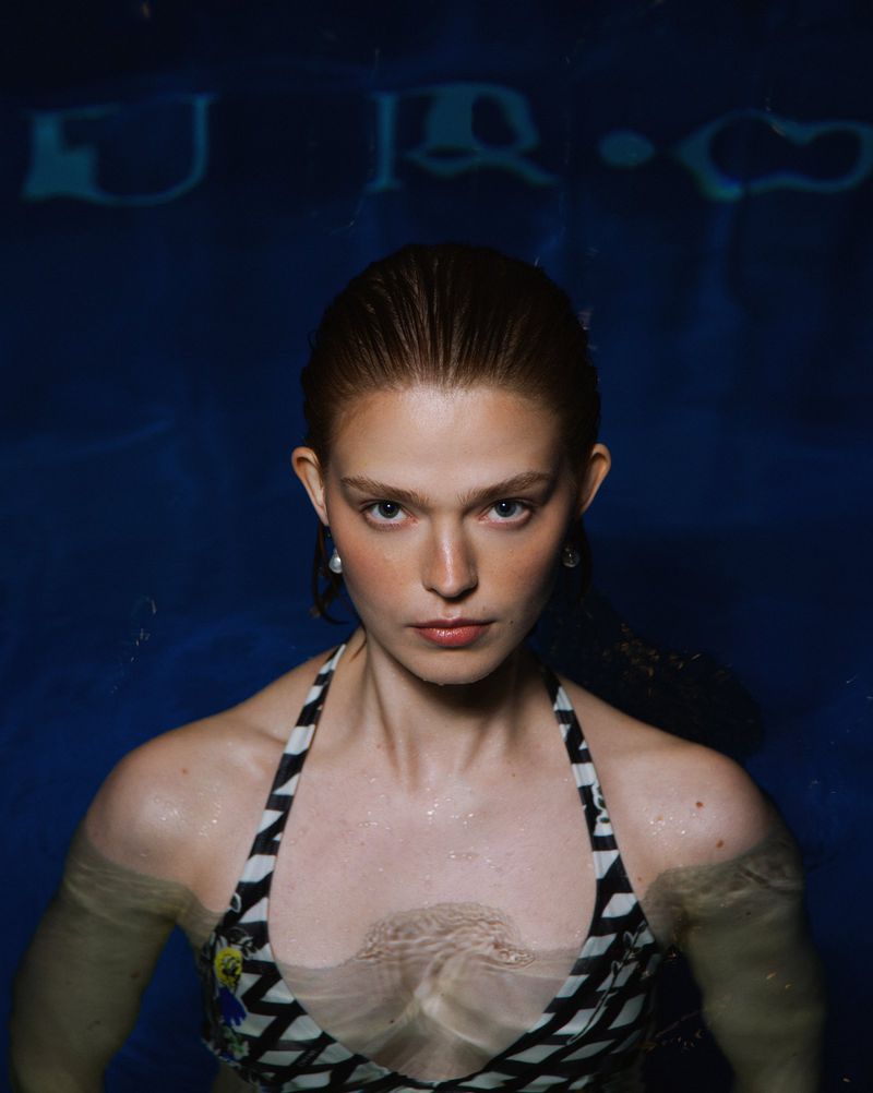 Attractive woman in a patterned swimsuit with wet hair emerging from blue water, looking directly at the camera with a serious expression.