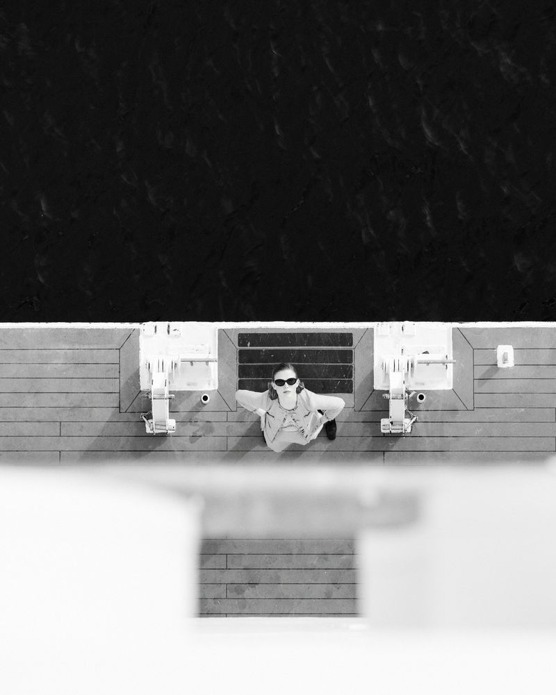 Overhead shot of a woman wearing sunglasses standing on a ship deck, looking up towards the camera, on a sunny day in black and white.