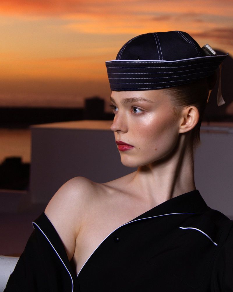 Profile of a young woman wearing a black sailor cap and a black top with white piping, against a sunset skyline background.
