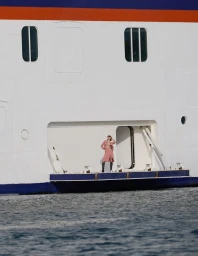 Model standing on a cruise ship platform in front of a massive white hull against blue water