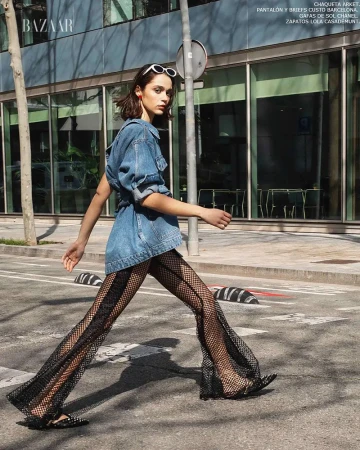 Fashion editorial shot of a woman walking across a street wearing a denim jacket and fishnet pants, styled with white sunglasses.