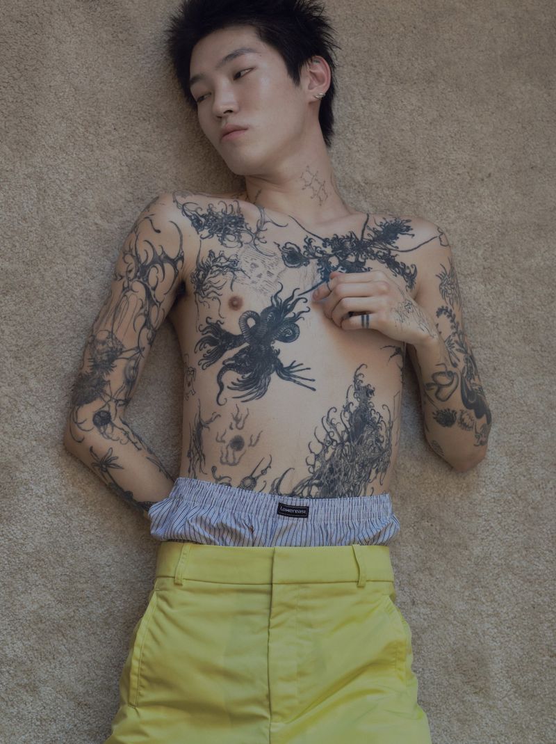Shirtless Asian male model with tattoos lies on carpet, wearing yellow pants and striped boxers. Indoor portrait with neutral background.