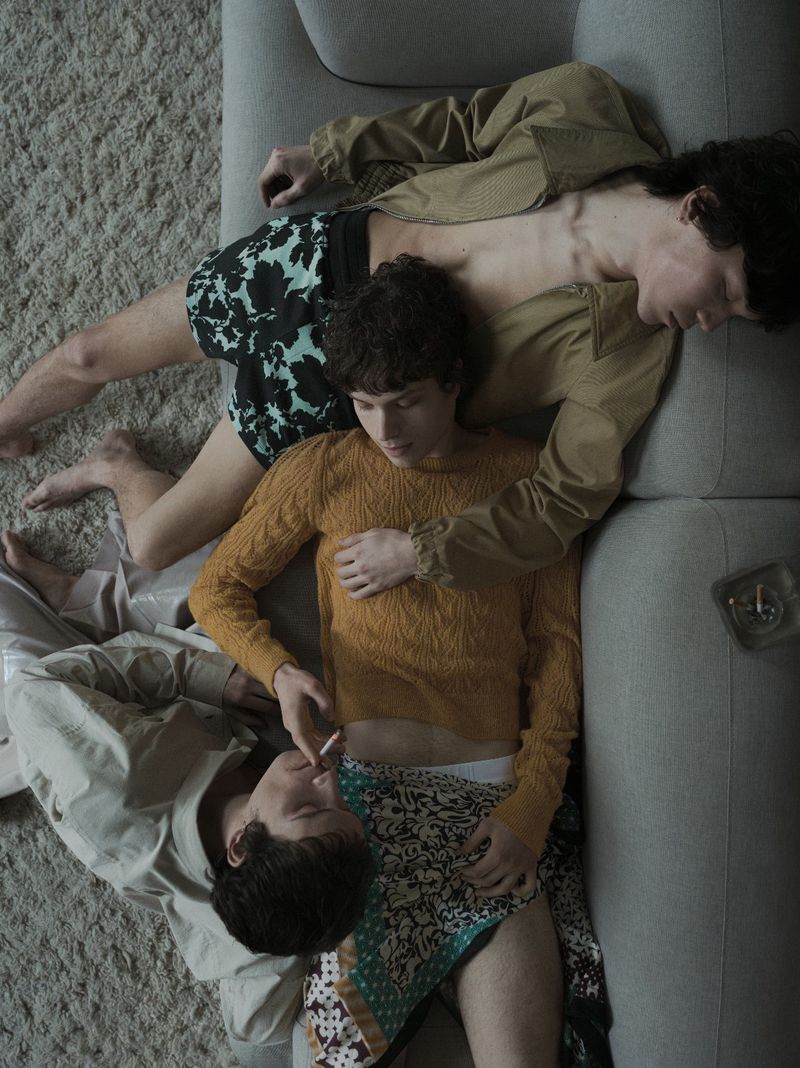Overhead shot of three young adult men lounging on a gray sofa in casual clothes and underwear, with a muted color palette and soft lighting.