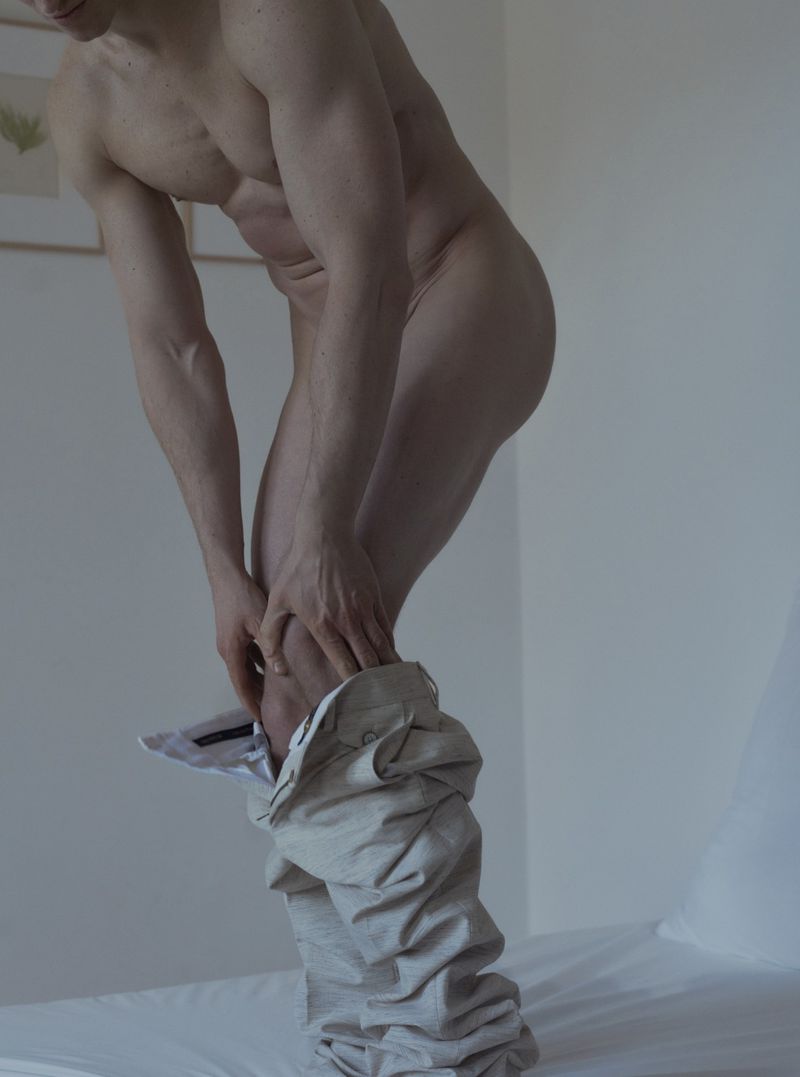 Nude adult male undressing indoors, holding pants on a bed in a high-angle portrait, showcasing muscular build and neutral expression.