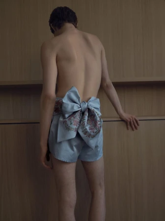 Back view of an adult with fair skin wearing blue shorts with a large bow, posing against a neutral, paneled wall in soft, diffused lighting.