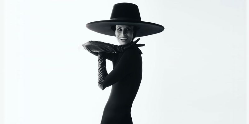 Monochrome studio portrait of a woman in a dark dress, large hat, and gloves, smiling with a pillow in her arms, bright white background.