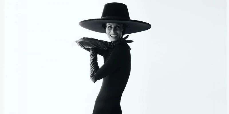 Monochrome studio portrait of a woman in a dark dress, large hat, and gloves, smiling with a pillow in her arms, bright white background.