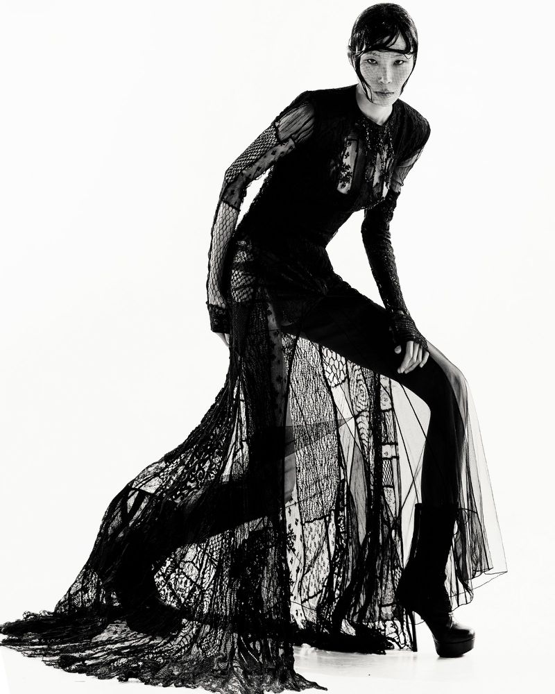 Monochrome fashion shot of an Asian woman in a long, patterned black dress and platform boots, striking a dynamic pose on a white backdrop.