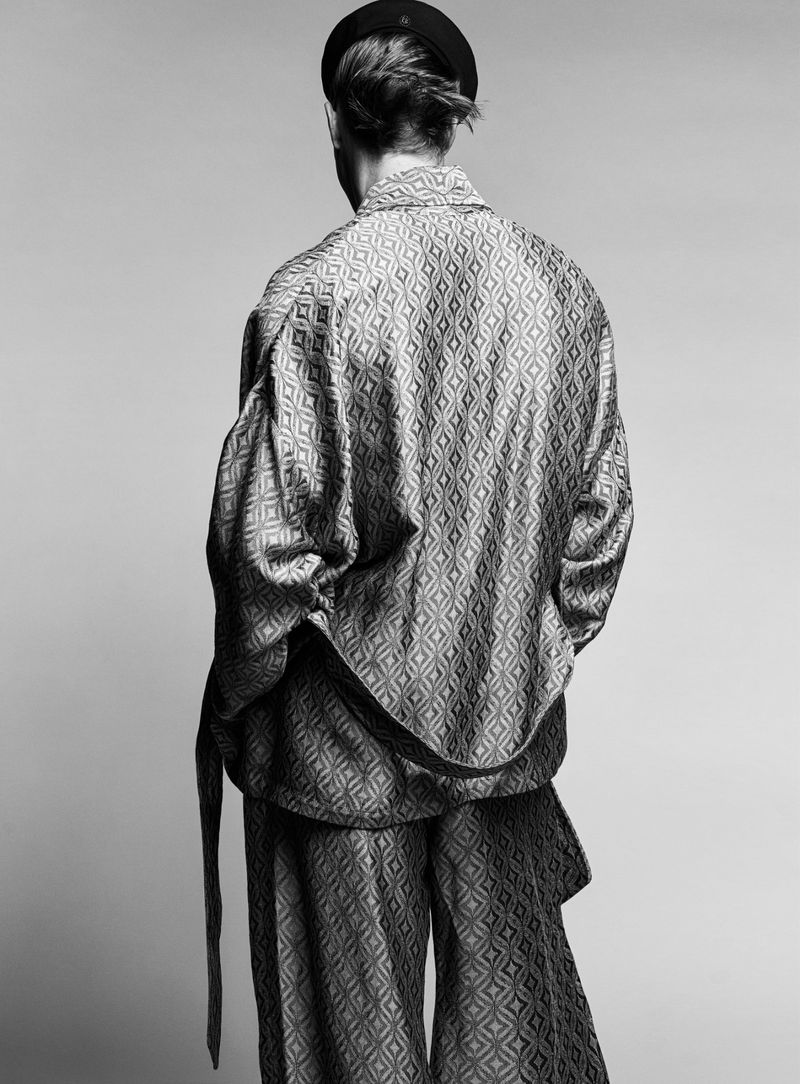 Back view of a person in a patterned shirt and pants, wearing a black beret, set against a neutral background.