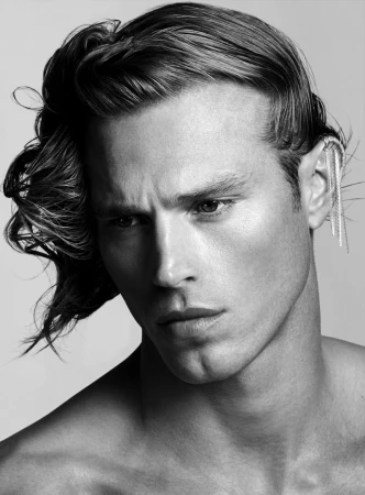 Striking portrait of a fair-skinned male model with wet-look wavy dark hair, showcasing an ornate ear cuff against a plain backdrop.