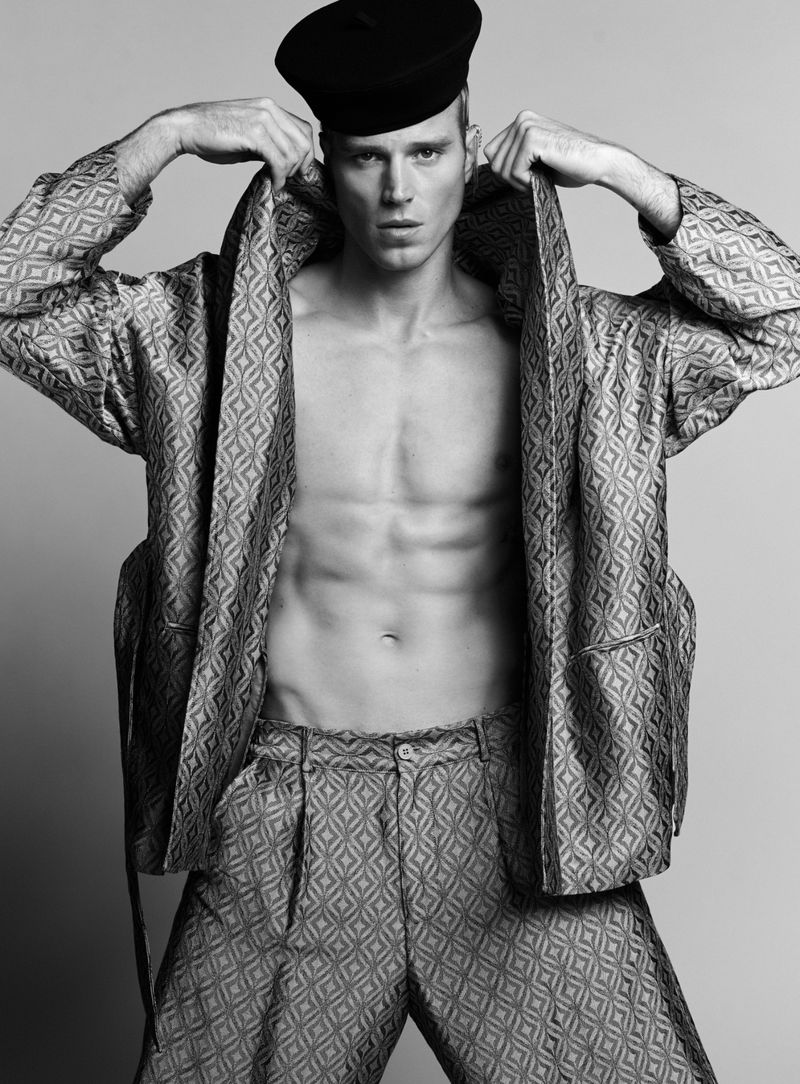 Male fashion model in a patterned jacket and pants with a black beret poses in a studio with a neutral background in a medium shot.