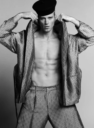 Male fashion model in a patterned jacket and pants with a black beret poses in a studio with a neutral background in a medium shot.