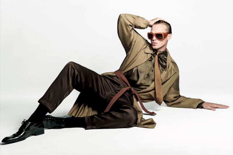 Adult man in a trench coat and sunglasses poses while lying down on a white surface in a fashion shot showing off his unique style.