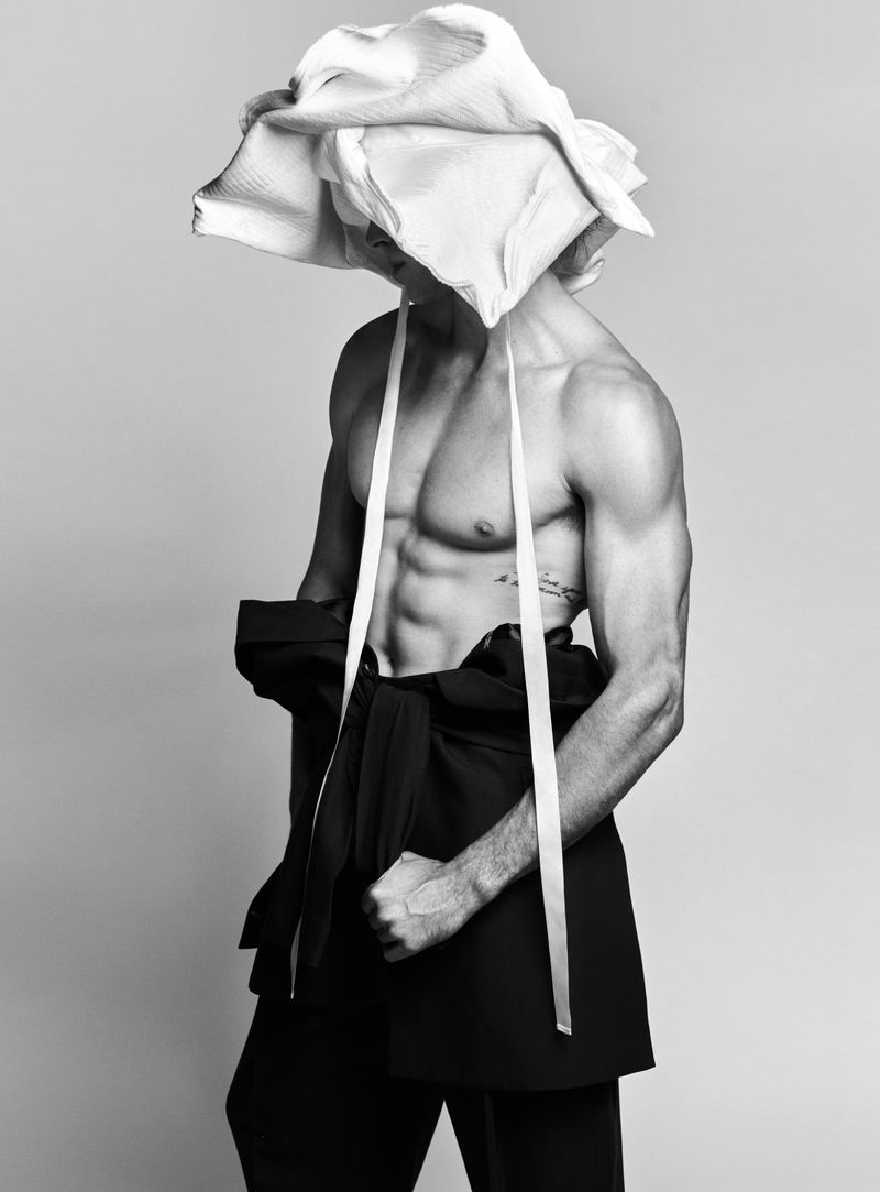 Black and white portrait of a muscular adult male wearing a flower hat, exposing his torso with a jacket draped on his waist.