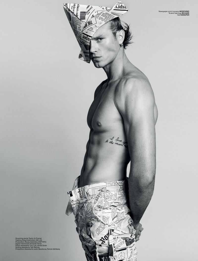 Shirtless male fashion model poses in newspaper-print pants and matching paper hat against a solid grey background in a studio shot.