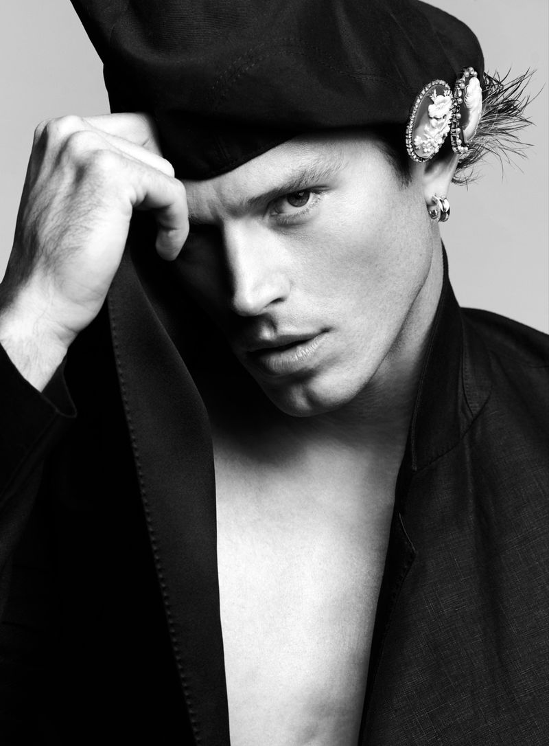 Portrait of a fair-skinned adult male with dark hair, wearing a beret and an open jacket, posing in front of a grey backdrop.