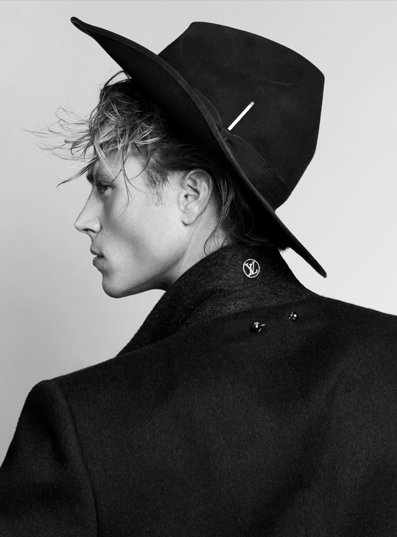 Black and white portrait of a male fashion model wearing a wide-brimmed hat and coat, posing in a photo studio with neutral lighting.