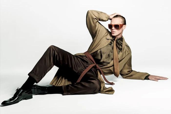 Matthew Noszka in a beige coat and brown trousers wearing sunglasses while posing on a seamless white floor
