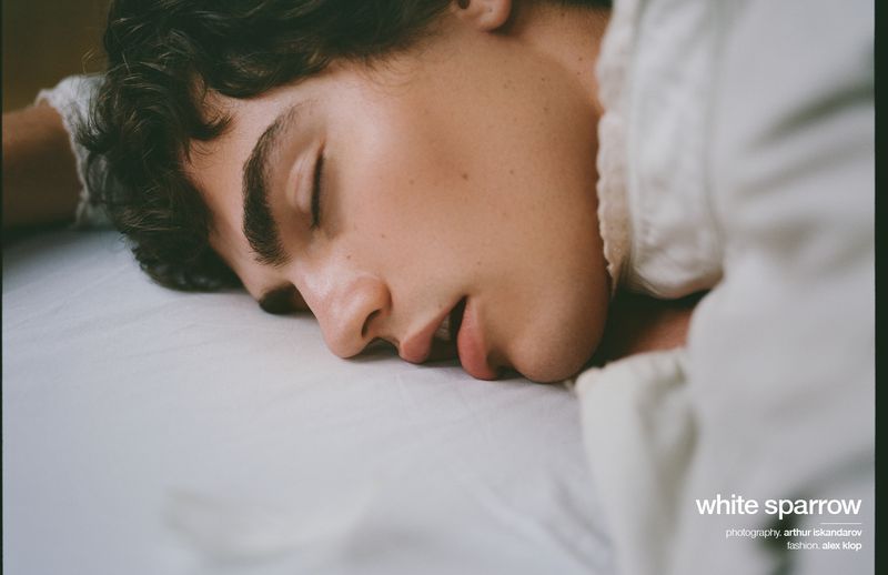 Close-up of a young male model with dark curly hair and closed eyes, lying on a crisp white sheet, illuminated by soft studio lighting.