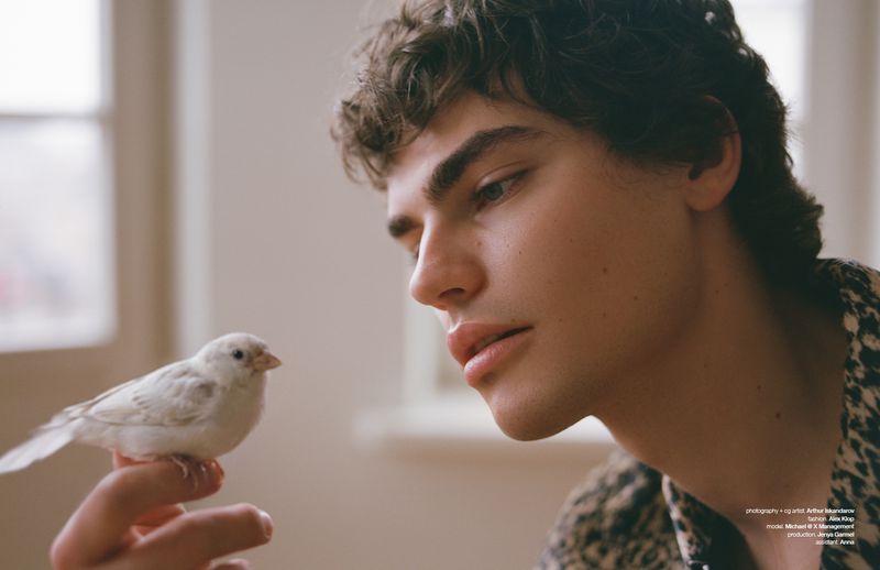 Portrait of a young man with curly dark hair holding a white bird on his finger, soft lighting, posing indoors.