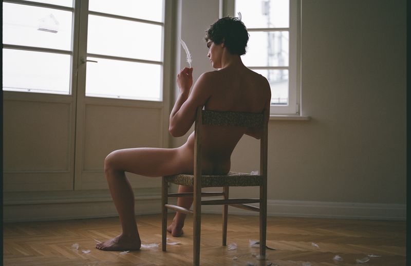 A person seated backwards on a wooden chair holding a feather in an interior room with a large window