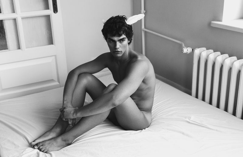 Nude male model with bunny ears sitting on a bed in a black and white photography studio setting with natural lighting.