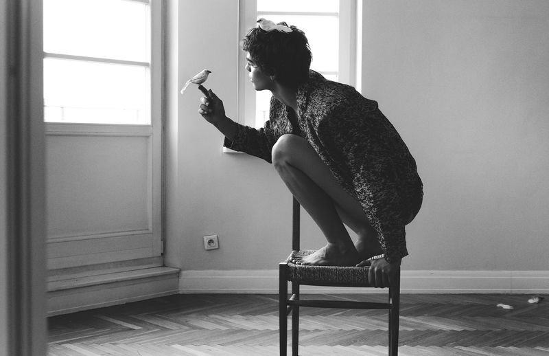 Monochrome image of person crouched on chair indoors, one bird on finger, another bird on head, neutral wall, and herringbone wood floor.