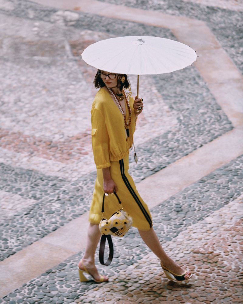 A woman in a yellow outfit walks on a cobblestone surface while holding a white parasol and a patterned purse with heels.