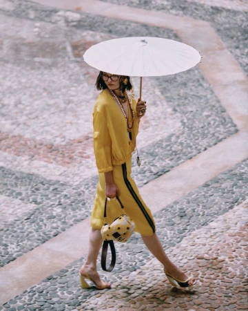 A woman in a yellow outfit walks on a cobblestone surface while holding a white parasol and a patterned purse with heels.