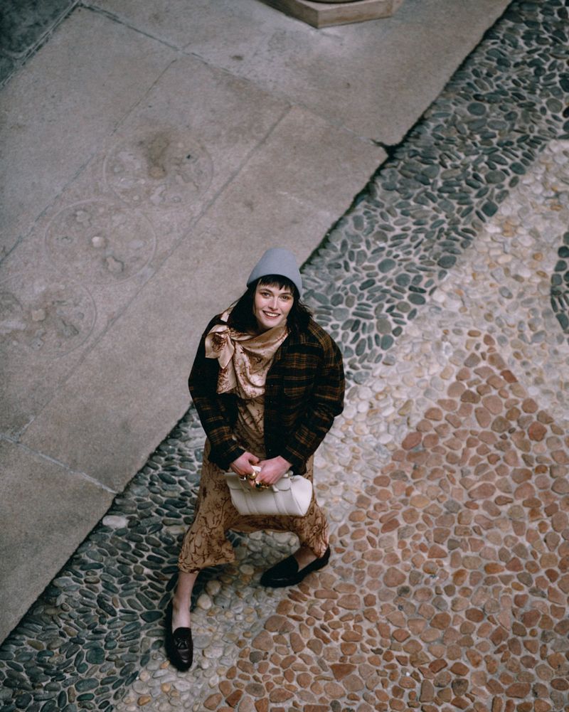 Overhead shot of a smiling adult woman in a patterned dress, coat, scarf, and hat holding a handbag against a stone mosaic.