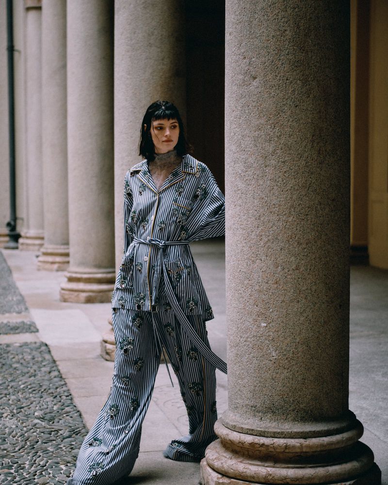 Fashion shot of a woman in a striped suit posing between stone columns, with a cobblestone floor and soft lighting in an outdoor setting.