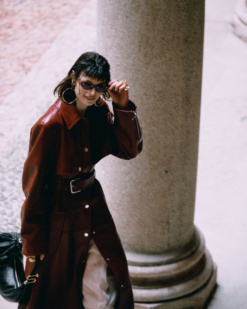 Woman in long red coat and sunglasses stands next to a stone column with handbag in an outdoor setting with light stone pavers.