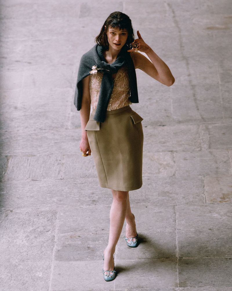Elegant adult female model in a beige top and skirt, light blue high heels, and a dark grey sweater draped around her shoulders.