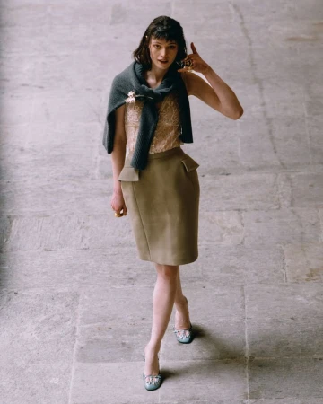 Elegant adult female model in a beige top and skirt, light blue high heels, and a dark grey sweater draped around her shoulders.
