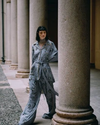 Model standing next to stone columns wearing a striped pajama-style matching shirt and pants set with embroidered details.