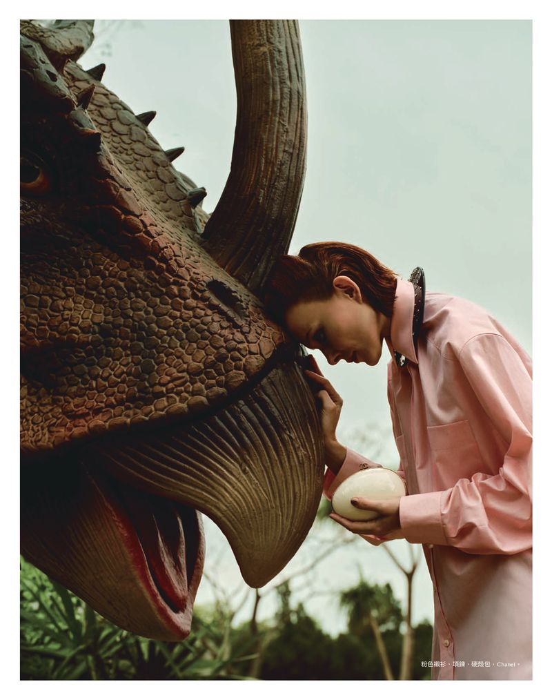 A woman in a pink shirt gently touches a dinosaur's head while holding an ostrich egg in an outdoor setting with muted colors and soft light.