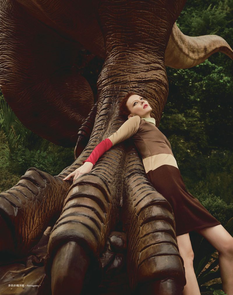 Woman leans against a dinosaur statue in an outdoor setting, wearing a beige and brown dress with red sleeves, looking up.