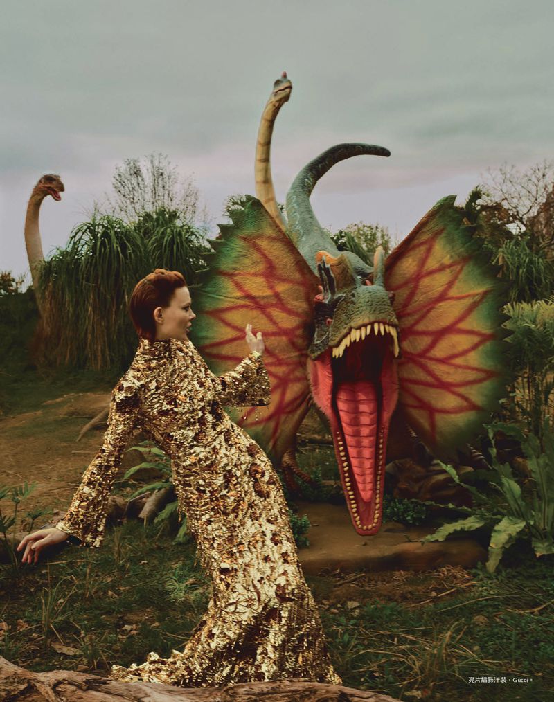 Woman in gold dress poses near dinosaur statues in a park-like setting with trees and sky, creating a whimsical and editorial image.