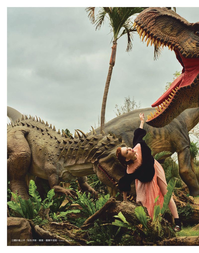 A woman in a pink dress poses with an open-mouthed dinosaur statue surrounded by lush greenery and a palm tree against a cloudy sky.