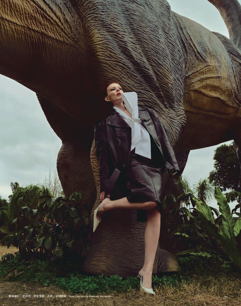 Adult woman in stylish clothing poses with a large dinosaur statue in an outdoor setting with natural lighting and lush greenery.