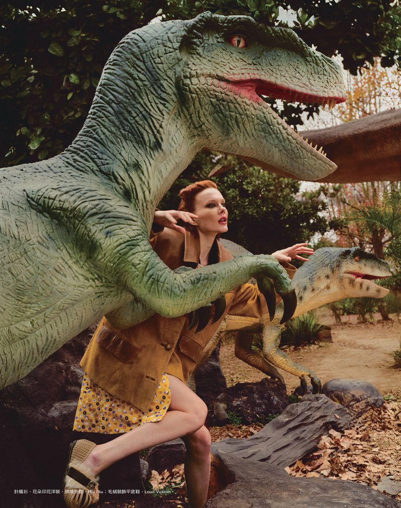 Woman poses dynamically with dinosaur statues outdoors, showcasing a floral dress and jacket in a staged editorial environment.